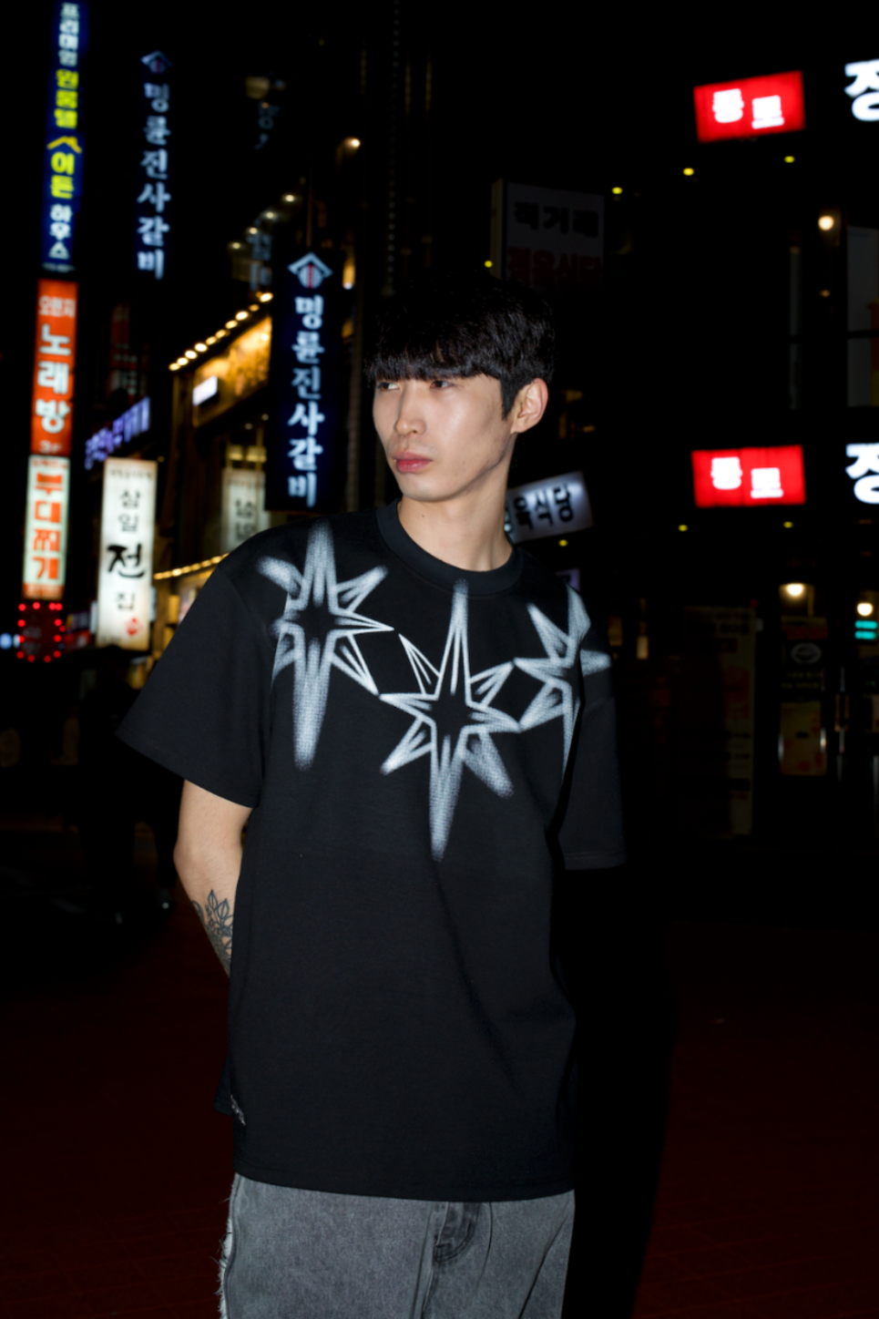 Person wearing a black t-shirt with white star design in an urban night setting with illuminated signs.